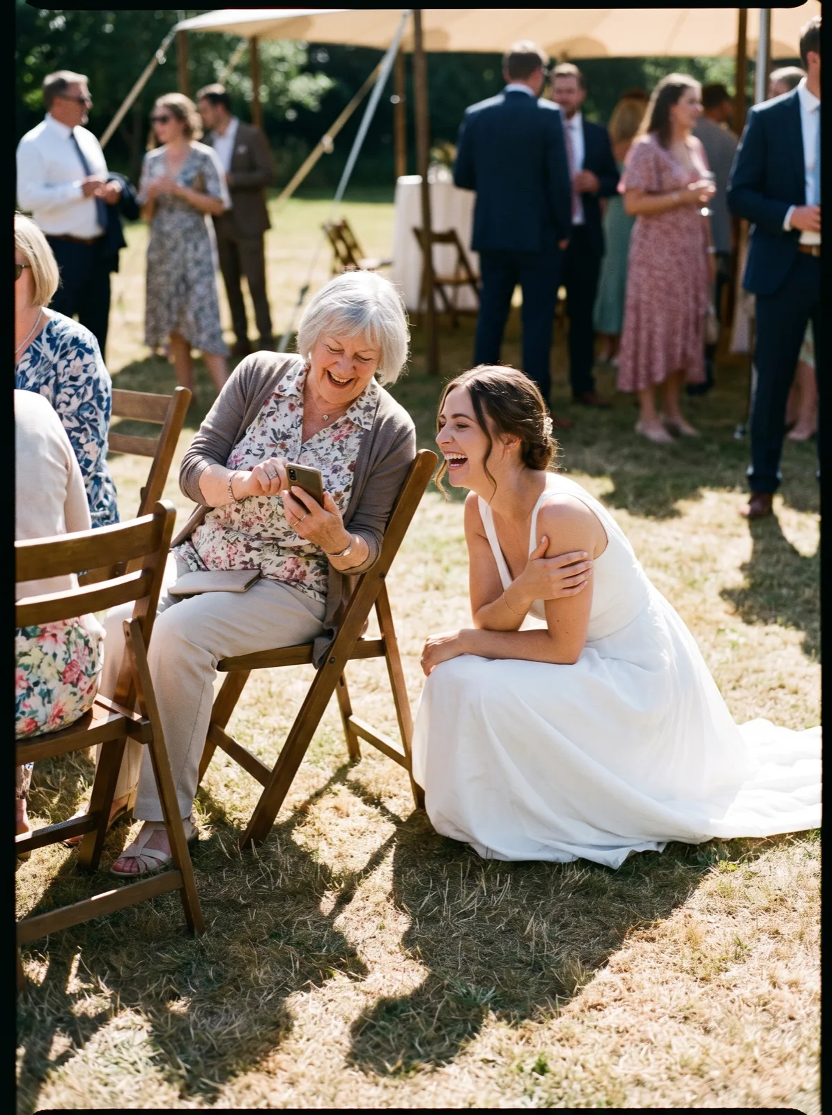 A bride crouching next to her grandmother at the wedding, both laughing at a photo on her phone